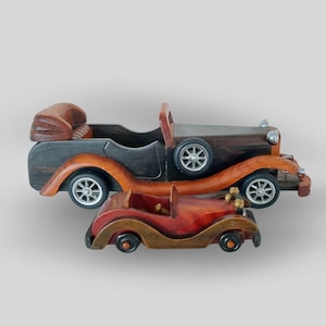 May include: Three wooden antique car models in varying sizes. The largest is a dark brown pickup truck with orange accents. The middle car is a dark brown convertible. The smallest is a red convertible with brown accents. All have silver wheels.