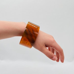 Puede incluir: Un brazalete ancho y translúcido en tonos ámbar y marrón. El brazalete grueso se lleva en la muñeca y tiene un aspecto veteado. El brazalete mide aproximadamente 6,35 cm de ancho.