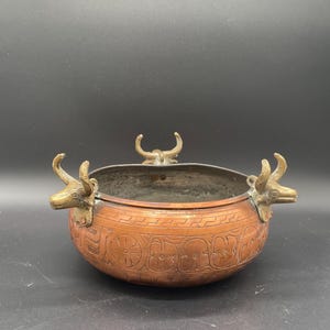 Peut inclure: Un bol en cuivre avec trois poignées en laiton en forme de tête de taureau. Le bol est orné d'un motif gravé complexe.