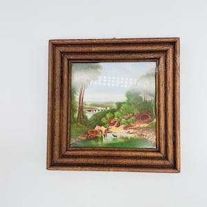 Puede incluir: Pintura de paisaje enmarcada con marco de madera. La obra de arte representa una escena serena con un río, un puente, árboles y figuras. La pintura tiene una estética vintage, con un enfoque en elementos naturales.