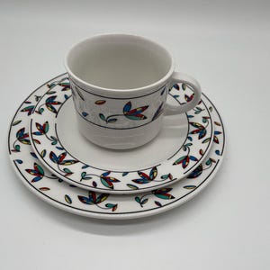 Può includere: Un set di tazza e piattino in ceramica bianca con un piattino abbinato. Il set presenta un bordo nero e un motivo ripetuto di foglie colorate in blu, rosso e giallo. La tazza ha un manico.