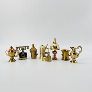 May include: A selection of miniature brass and copper ornaments, including a teapot, telephone, watering can, iron, lamp, mortar, and jug. The items are displayed on a white surface, highlighting their intricate details and vintage style.