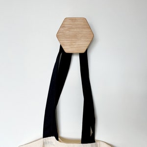 May include: A wooden hexagonal hook with a tote bag hanging from it. The hook is mounted on a white wall. The tote bag has black straps and a cream-colored canvas body.