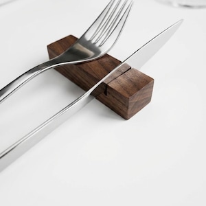 Puede incluir: Un reposacubiertos rectangular de madera marrón oscuro con una ranura estrecha en la parte superior. Un tenedor de acero inoxidable pulido y un cuchillo de mesa a juego están colocados descansando en la ranura del bloque de madera.