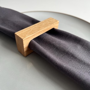 May include: A dark gray linen napkin is held by a rectangular wooden napkin ring. The napkin ring is made of light-colored wood and has a simple, modern design. The napkin is resting on a light gray plate.