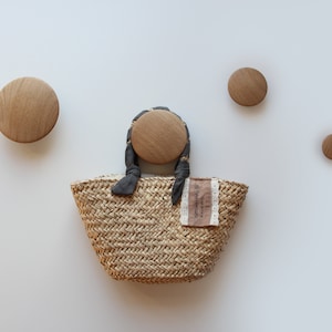 Peut inclure: Un petit sac cabas en paille tressée avec une anse en ruban noir et des accents en bois. Le sac a une petite étiquette blanche avec le texte "el taller de la abuela" dessus.