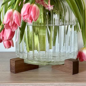 May include: A clear glass vase with pink and white tulips. The vase sits on two small, dark wooden stands. The tulips have green stems and leaves. The vase has a ribbed design and is filled with water.