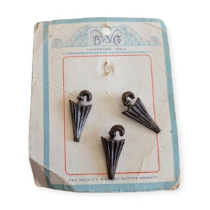 May include: Three dark brown umbrella-shaped buttons attached to a vintage off-white cardboard display card. The card features blue decorative borders and text that reads B.W.C. Muscatine Iowa and The World's Biggest Button Market.
