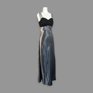 May include: A formal, floor-length gown on a mannequin. The bodice is black with a sweetheart neckline and thin straps. The skirt is a shimmering, silvery-gray material that flows to the floor. The dress is displayed against a gray background.