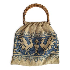 May include: A beige drawstring bag with a bamboo handle. The bag features a blue embroidered design of two birds flanking a central vase. The handle is made of curved bamboo.
