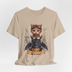 May include: A beige t-shirt with a cartoon image of a cat wearing goggles riding a black dragon. The text "Cadet Broccoli" is printed below the image.