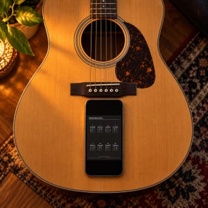 Pode incluir: Uma guitarra ac&uacute;stica com corpo de madeira clara, ponte escura e escudo de tartaruga. Um smartphone est&aacute; colocado sobre o corpo da guitarra, exibindo um ecr&atilde; com o texto Essential Open Chords e uma grelha de oito diagramas de acordes.