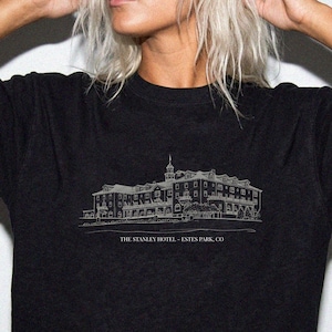 May include: A black short-sleeved t-shirt featuring a white line-art illustration of The Stanley Hotel. Below the building graphic, the text reads THE STANLEY HOTEL - ESTES PARK, CO in a simple, clean, sans-serif font centered on the chest.