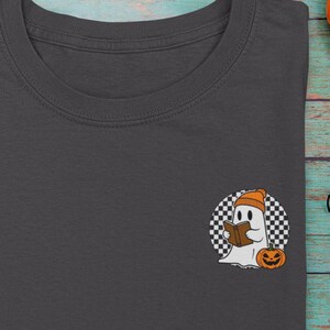 May include: Dark gray t-shirt featuring a Halloween-themed graphic. The graphic includes a ghost wearing a beanie and holding a book, set against a checkered background with a pumpkin. The shirt is folded on a teal wooden surface with Halloween decorations.