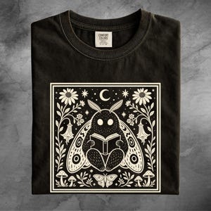 May include: A black t-shirt featuring a white graphic print of a moth reading an open book. The design is framed by flowers, stars, a crescent moon, and smaller mushrooms. The inner neck label displays the text Comfort Colors.