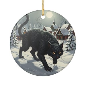 May include: A round ceramic ornament featuring a black cat with intricate silver swirl patterns on its fur, yellow eyes, and a raised tail. The cat stands in a snowy village scene with pine trees, small houses, and a full moon in the background.