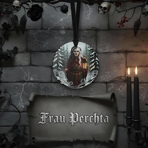 May include: A circular hanging ornament featuring an illustration of a figure in a dark red cloak holding a glowing lantern amidst a snowy forest. Below the ornament is a scroll-shaped sign with the text Frau Perchta written in black gothic font.