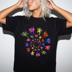 May include: A black short-sleeved t-shirt featuring a central graphic print of numerous small, colorful cartoon-like figures with wings arranged in a swirling spiral pattern. The figures are rendered in bright shades of red, blue, green, yellow, and purple.