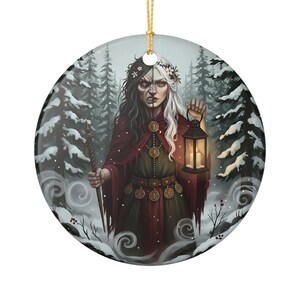May include: A round ceramic ornament featuring a split-faced figure with dark and light hair, wearing a red cloak and dark tunic. The figure holds a staff and a lit lantern in a snowy forest setting with pine trees. A gold string hangs from the top.