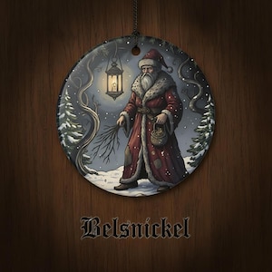 May include: A round ceramic ornament featuring a detailed illustration of Belsnickel in a long red fur-trimmed coat, holding a lantern and a bundle of sticks in a snowy forest. The word Belsnickel is printed in a black gothic font below the ornament.