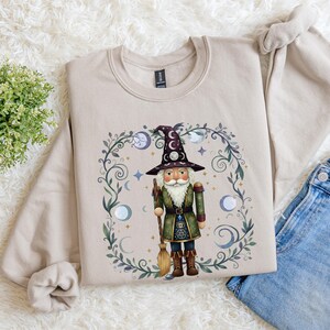 May include: Beige sweatshirt featuring a whimsical illustration of a wizard nutcracker. The design includes a tall hat, broom, and a decorative border with moons and stars. The sweatshirt is paired with blue jeans.