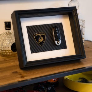 Puede incluir: Una imagen enmarcada en negro con un fondo de fibra de carbono. La imagen presenta un logotipo dorado de Lamborghini y una llave de coche plateada.