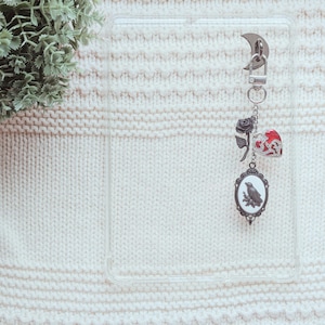 May include: A silver keychain with a crescent moon clasp, featuring charms of a black rose, a red heart, and an oval pendant with a raven illustration. The charms hang from a silver chain. The background is a white knitted fabric.
