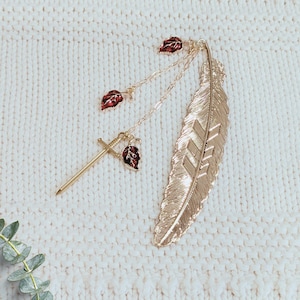 May include: Gold-coloured feather bookmark with a sword and leaf charm. The bookmark features a detailed feather design and is accented with small red and gold leaves. A unique accessory for book lovers.