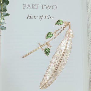 Puede incluir: Marcapáginas de metal dorado con forma de pluma, con un dije de espada y hojas verdes. El marcapáginas está sobre una página de libro con el texto "PART TWO Heir of Fire". Hojas de eucalipto en el fondo.