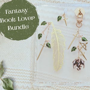 Puede incluir: Una funda de acrílico transparente con colgantes de temática fantástica. Los colgantes incluyen un marcapáginas de pluma dorada, espadas, un arco y flechas y un dragón. El texto "Fantasy Book Lover Bundle" está en un círculo verde.
