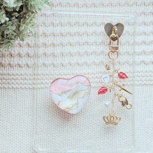 May include: Clear phone case with a heart-shaped pop socket featuring pink, gold, and white marbling. A gold charm with a heart, crown, sword, and red leaf accents hangs from the case. The case rests on a cream-colored knitted surface.