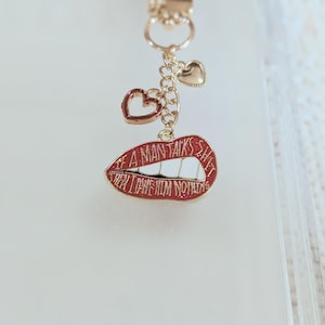 May include: A gold-colored keychain charm with a red lips pendant that reads "IF A MAN TALKS SHIT THEN I OWE HIM NOTHING". The charm also features two heart-shaped pendants.