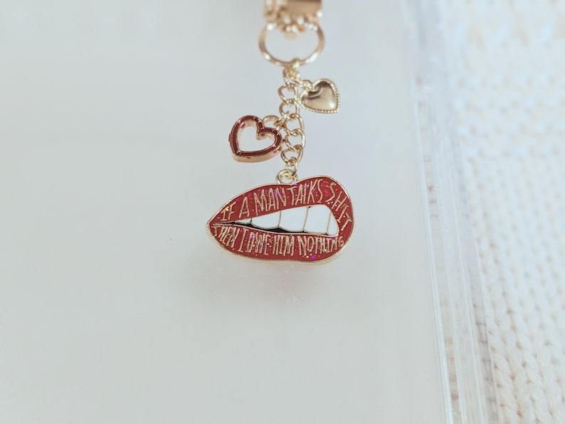 May include: A gold-colored keychain charm with a red lips pendant that reads "IF A MAN TALKS SHIT THEN I OWE HIM NOTHING". The charm also features two heart-shaped pendants.