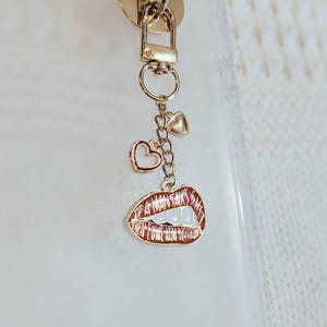 May include: Gold-toned keychain with a lobster clasp, two heart charms, and a lip-shaped charm. The lip charm is red with white teeth and the words "I WANT IT ALL" in white.