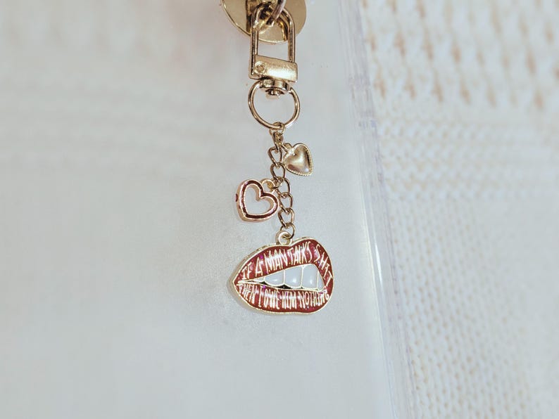 May include: Gold-toned keychain with a lobster clasp, two heart charms, and a lip-shaped charm. The lip charm is red with white teeth and the words "I WANT IT ALL" in white.