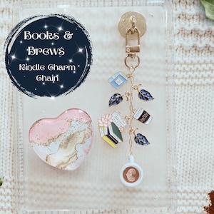 May include: A clear phone case with a heart-shaped pop socket and a charm chain. The charm chain features book, coffee cup, and leaf charms. The pop socket is pink, gold, and white. The text "Books & Brews Kindle Charm Chain" is in a dark blue circle.