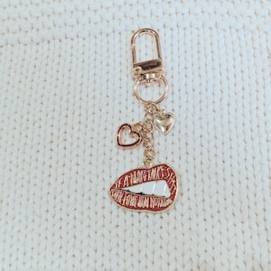 May include: A gold-colored keychain with a lobster clasp, three heart charms, and a red lips charm. The lips charm has the text "A MAN TALKS HAS HER FROM NOTHING" in white lettering.