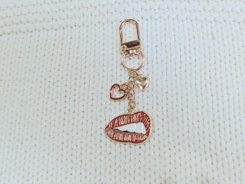 May include: A gold-colored keychain with a lobster clasp, three heart charms, and a red lips charm. The lips charm has the text "A MAN TALKS HAS HER FROM NOTHING" in white lettering.