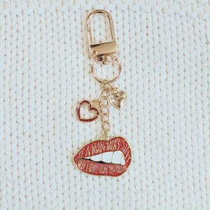 May include: Gold-tone keychain with a lobster clasp, small heart charms, and a red lips charm. The lips charm has white teeth and the text "A MAN'S KISS IS THE BEST SHIT". The keychain is on a white knitted surface.