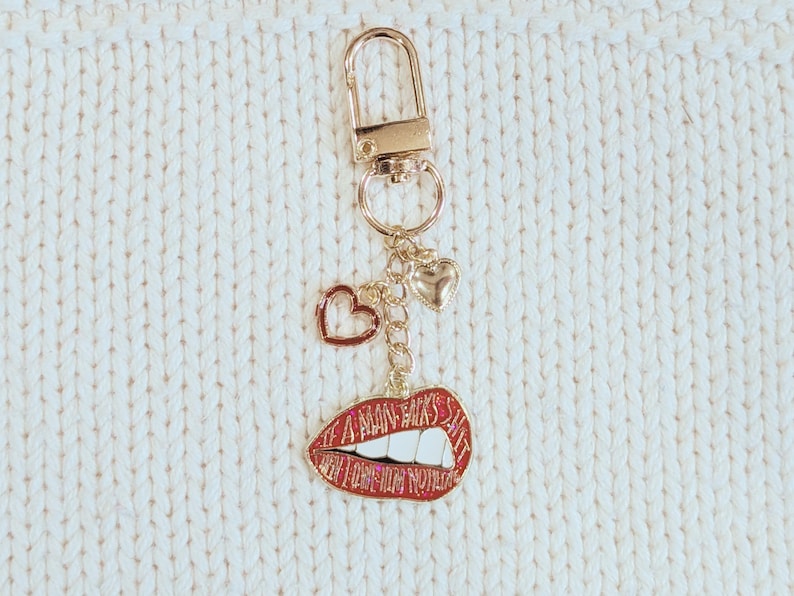 May include: Gold-tone keychain with a lobster clasp, small heart charms, and a red lips charm. The lips charm has white teeth and the text "A MAN'S KISS IS THE BEST SHIT". The keychain is on a white knitted surface.