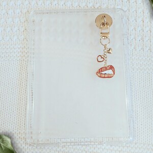May include: Clear phone case with a gold-colored charm. The charm features a small heart, a camera, and a red lips pendant. The case is rectangular and has a gold-colored attachment at the top.