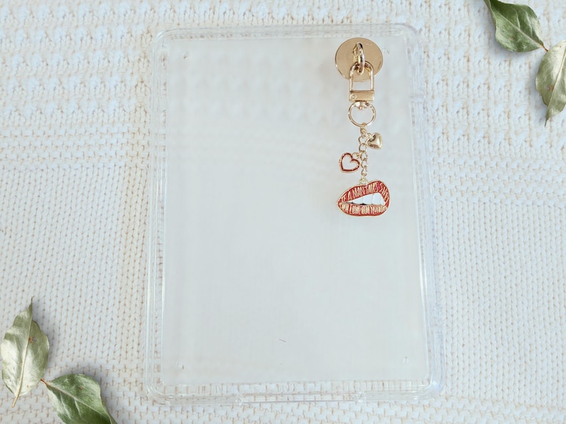 May include: Clear phone case with a gold-colored charm. The charm features a small heart, a camera, and a red lips pendant. The case is rectangular and has a gold-colored attachment at the top.