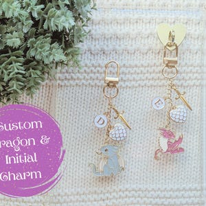 May include: Two keychains with dragon and initial charms. One keychain has a blue dragon charm and the letter "D". The other keychain has a pink dragon charm and the letter "E". Both keychains have gold-colored hardware and decorative charms.