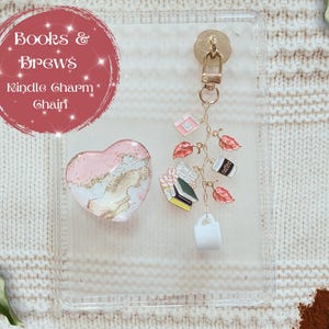 May include: A clear phone case with a gold-colored charm chain featuring book, coffee cup, and leaf charms. A pink and gold heart-shaped popsocket and the text "Books & Brews Kindle Charm Chain" are also visible.