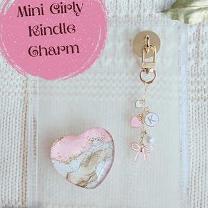 Puede incluir: Un charm de Kindle en forma de corazón rosa y dorado con un cierre dorado y un charm con un lazo rosa, corazones y la letra "K". El texto "Mini Girly Kindle Charm" está en un círculo rosa.