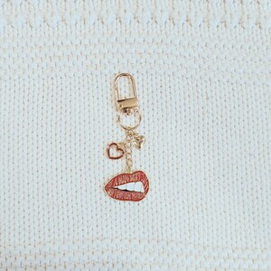 May include: A gold-colored keychain with a lobster clasp, featuring a charm with red lips, a heart, and a small star. The lips charm has a red enamel finish with white teeth. The keychain hangs against a white, textured background.