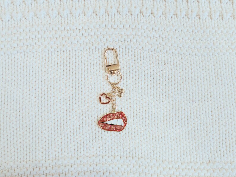 May include: A gold-colored keychain with a lobster clasp, featuring a charm with red lips, a heart, and a small star. The lips charm has a red enamel finish with white teeth. The keychain hangs against a white, textured background.