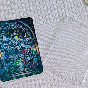 Può includere: Una custodia rettangolare per telefono con un design olografico colorato di un paesaggio montano, stelle e una luna crescente. Accanto, una custodia trasparente.