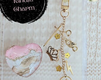 Könnte beinhalten: Eine transparente Handyhülle mit einem herzförmigen Anhänger in Rosa und Gold sowie einer goldfarbenen Charm-Kette. Die Kette zeigt ein Schwert, einen Schmetterling, Blumen, einen Blitz und eine Wolfssilhouette. Der Text "Fantasy Kindle Charm" befindet sich oben links.