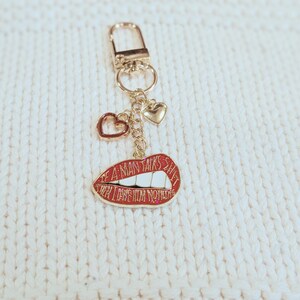 May include: Gold-tone keychain with a red lips charm that reads "IF A MAN TALKS SHIT THEN LOVE HIM NOTHIN". The keychain also features two heart charms. The background is a white, textured surface.